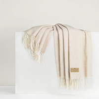 The Lands Downunder Juno Cashmere Throw is a soft, cream-colored luxury blanket with fringe edges and a brown label on one corner, elegantly draped over a white rectangular block.