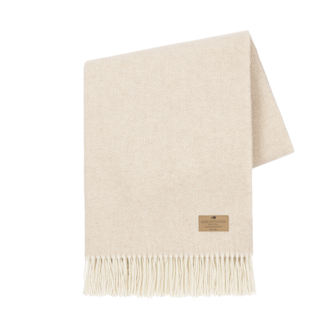 The Juno Cashmere Throw by Lands Downunder is a cream lambswool blanket with a subtle pattern, fringed edges, and a small brown label—an elegant touch of luxury for any space.