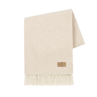 The Juno Cashmere Throw by Lands Downunder is a cream lambswool blanket with a subtle pattern, fringed edges, and a small brown label—an elegant touch of luxury for any space.