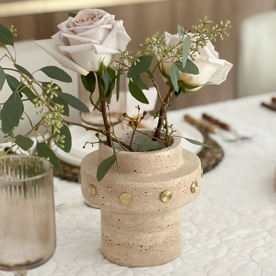 The BT-Shalom Tulsa Vase with gold studs holds pale pink roses and greenery on a white textured tablecloth, beside a clear ribbed glass and a set dining table in the background.