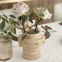 The BT-Shalom Tulsa Vase with gold studs holds pale pink roses and greenery on a white textured tablecloth, beside a clear ribbed glass and a set dining table in the background.