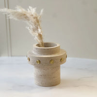 The Tulsa Vase by BT-Shalom, featuring modern travertine with gold rivet details, rests on a white surface and holds a single stem of dried pampas grass against a soft, minimal background that highlights its unique design.