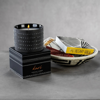 A Kari Candle by Zodax with a tobacco vanilla scent in a textured ceramic holder rests on its labeled box. Nearby, a woven bowl holds two books and matches on a gray surface, bringing cozy fragrance to the space.