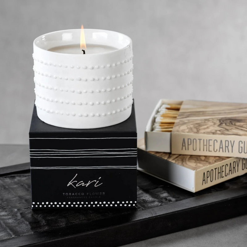 A white textured Zodax Kari Candle sits on a black box next to a partially open matchbox and two books titled APOTHECARY GUIDE, all arranged on a dark tray against a gray background.