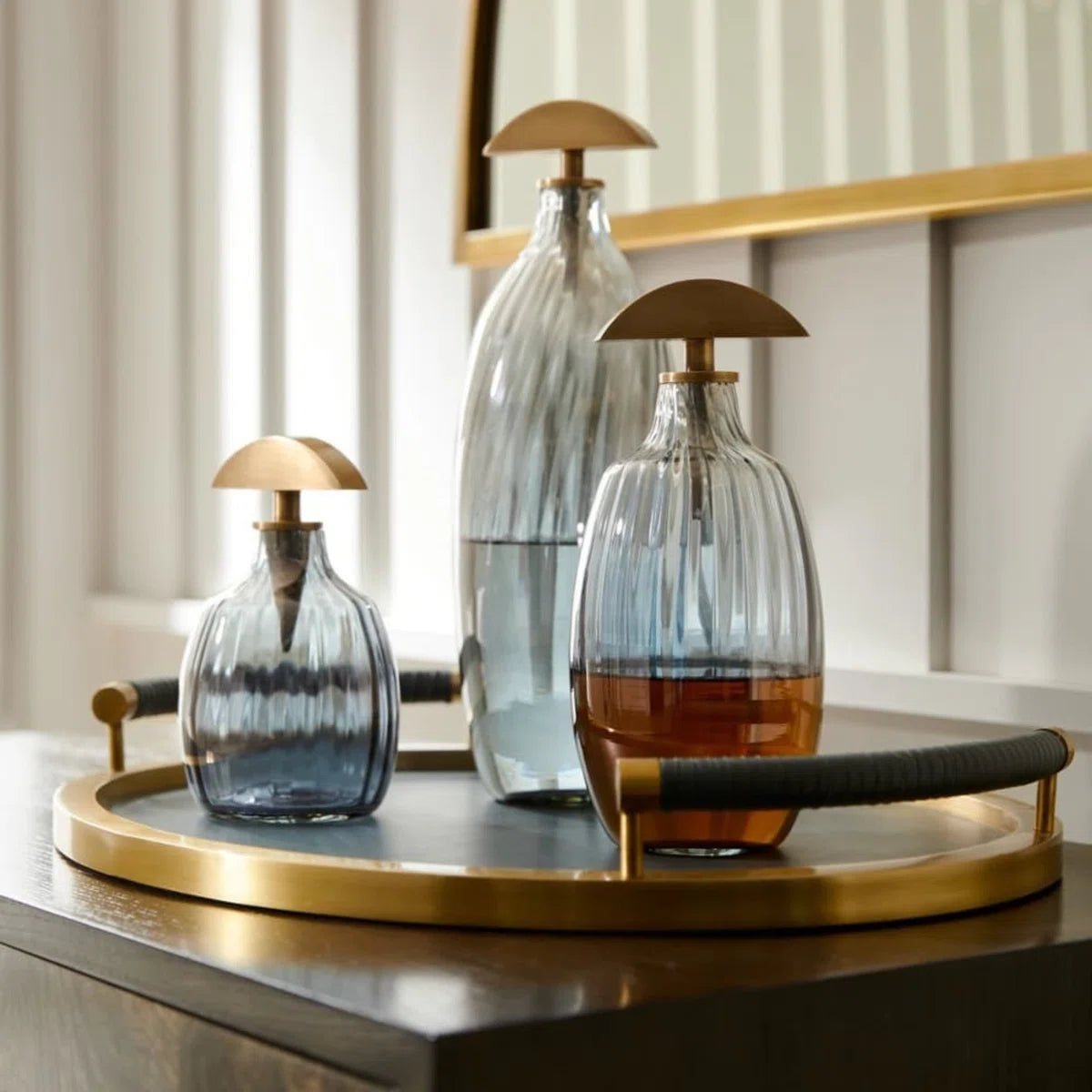 Three Karis Decanter pieces by Arteriors, each with ribbed optic glass and gold dome stoppers, rest on a round antique brass tray atop wood, backed by a mirror and white paneled wall.