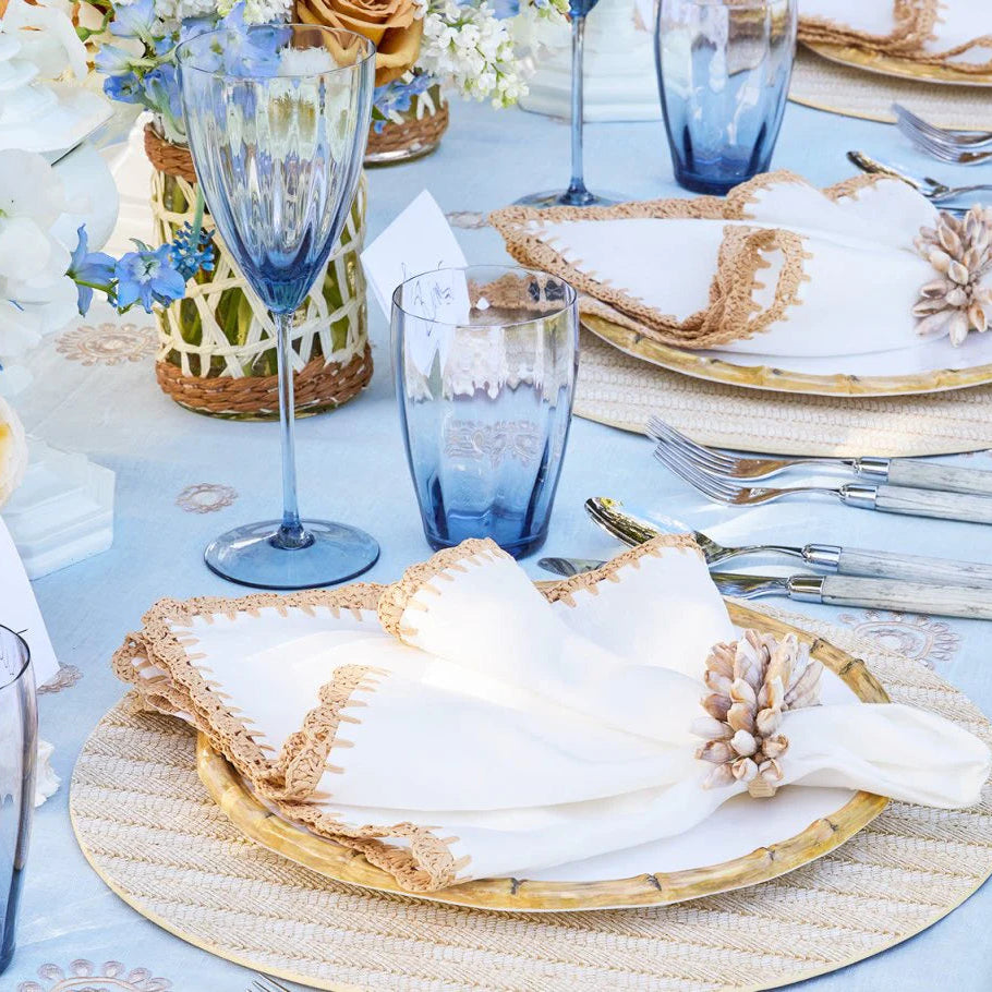 A close-up of an elegant beach-inspired table set with blue glasses, gold-rimmed plates, white napkins with tan trim and Kim Seybert’s Shell Fringe Napkin Ring Set of 4, atop a light blue floral tablecloth.