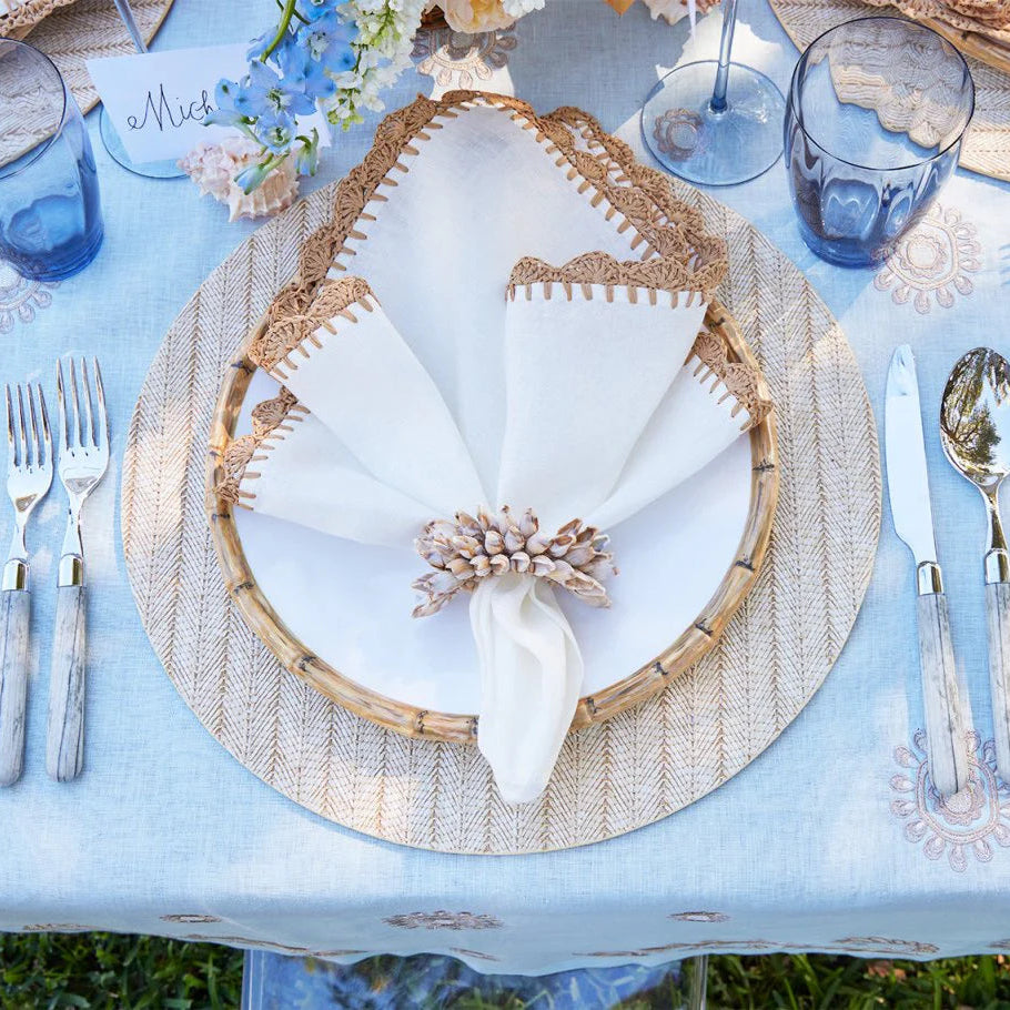 A beach-inspired table setting features a Kim Seybert Shell Fringe Napkin Ring Set of 4 with beaded shells, paired with a round placemat, white plate, folded napkin, blue glasses, silver cutlery, and a floral centerpiece on a pale blue tablecloth.