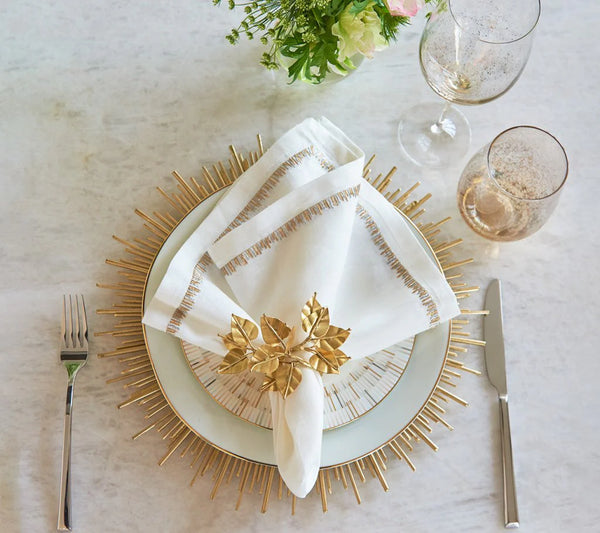 An elegant formal table setting features Kim Seybert's Radiate Charger in Gold, white plates, a folded white napkin with gold trim in a gold leaf ring, two wine glasses, fork on the left, knife on the right, and flowers as décor.