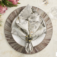 A beautifully set table features a grey floral napkin, Kim Seybert’s Bonton Napkin Ring Set of 4 in Grey & Gold, elegant plates, and a wooden charger, with pink protea flowers and greenery adding vibrant color.