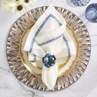 A stylish table features a gold plate on a patterned charger, a white napkin with blue embroidery, and a Kim Seybert Bonton Napkin Ring Set of 4-Navy & Gold, accented by marble, purple glassware, and white flowers.
