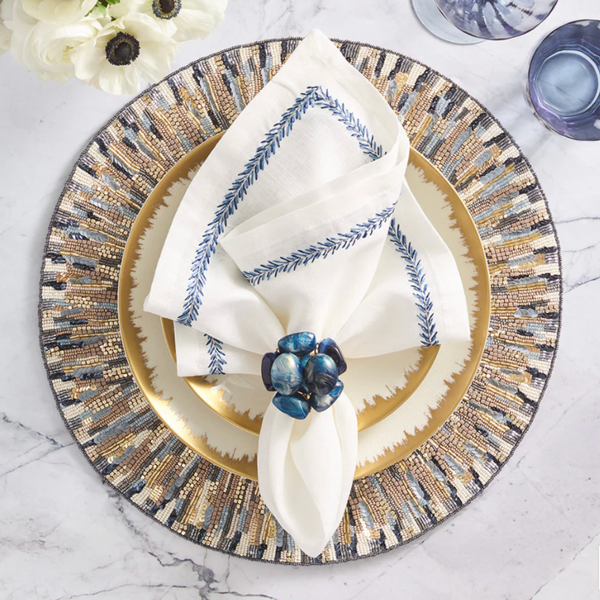 A stylish table features a gold plate on a patterned charger, a white napkin with blue embroidery, and a Kim Seybert Bonton Napkin Ring Set of 4-Navy & Gold, accented by marble, purple glassware, and white flowers.