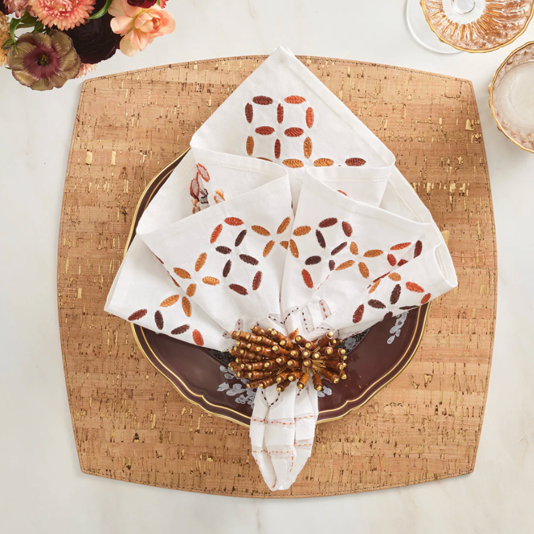 A square cork placemat holds a dark brown plate with gold trim, topped by a white napkin with an orange and brown geometric pattern, secured with a Kim Seybert Royal Palm Napkin Ring Set of 4. Tropical table decor and glassware rest on the marble surface.