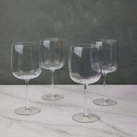 Four empty, clear Borough Barware glasses by LSA are arranged on a white marble surface with a gray textured background. Their reflections add elegance, making this Set of 4 a stylish addition to any barware collection.