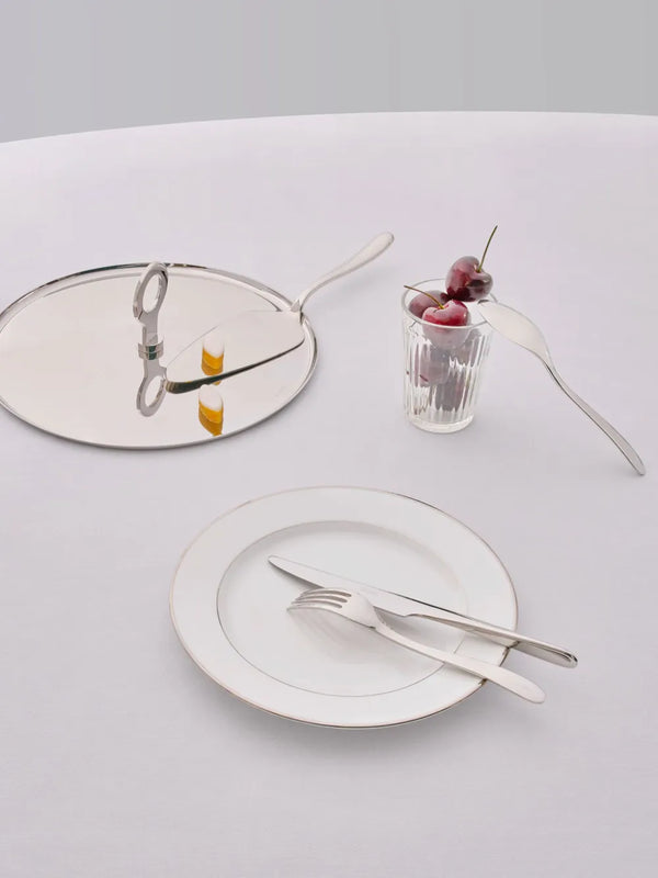 A L’Âme de Christofle Stainless Steel Serving Fork and cake server rest on a mirrored tray, accompanied by a plate, silverware, and a glass of cherries on a white tablecloth.