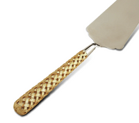 The L'Objet Braided Cake Server features a flat, triangular metal blade and a light brown handle with an elegant crisscross design. Part of the Braid serveware collection, its basket-style handle adds sophisticated detail.