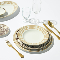 A set of Ginori 1735 Labirinto Dinnerware - Cashemire, adorned with gold Greek key patterns, is displayed alongside two gold utensils and two clear glasses on a white surface.