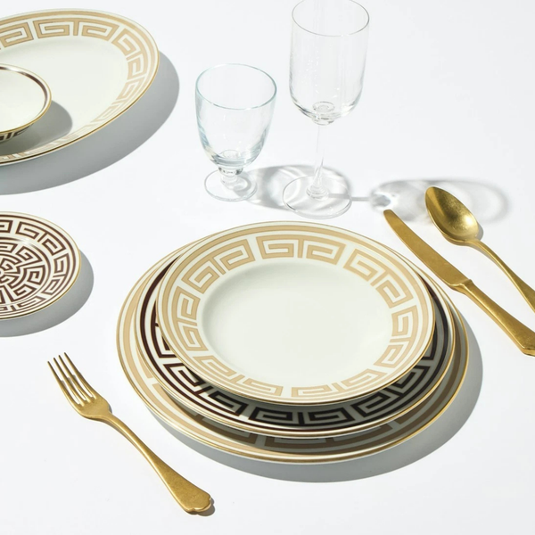 A set of Ginori 1735 Labirinto Dinnerware - Cashemire, adorned with gold Greek key patterns, is displayed alongside two gold utensils and two clear glasses on a white surface.