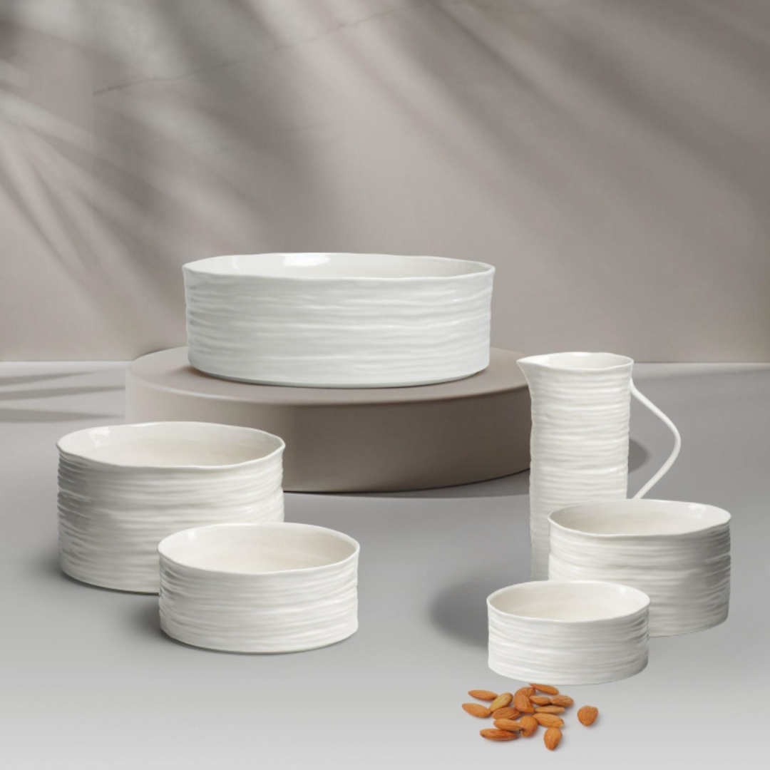The PAMPA BAY Le Blanc Bowl set features white porcelain dishes and bowls with a textured, wavy surface. Oven and dishwasher safe, they are arranged on a light background with almonds in the foreground and soft shadows behind.