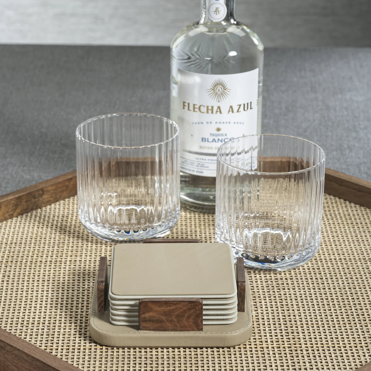 A bottle of Flecha Azul tequila, two ribbed glass tumblers, and a stack of Zodax Leather & Veneer Coaster Set in Cappuccino rest on a woven beige tray with wooden edges.