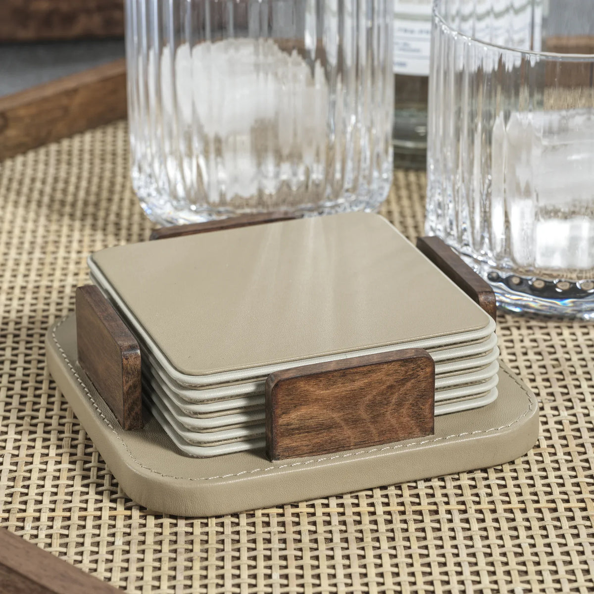 The Zodax Leather & Veneer Coaster Set in Cappuccino rests in its wooden holder on a woven tray, surrounded by clear textured glass tumblers.