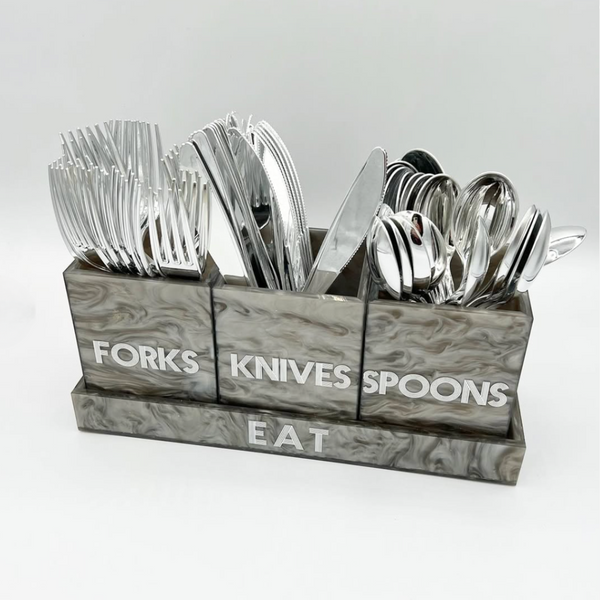 The Eye Candy Let's Eat Silverware Caddy in Grey Marble features three labeled containers for forks, knives, and spoons with silver utensils, set in a matching acrylic "EAT" tray—an elegant choice for any modern table.