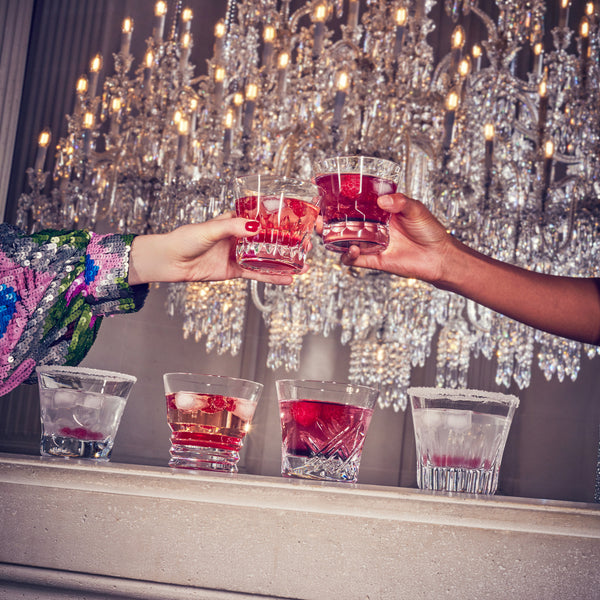 Two hands clink cut crystal glasses filled with red drinks before a sparkling chandelier, while four more cocktails in the Baccarat Everyday Tumblers Set of 6 line a marble counter below.