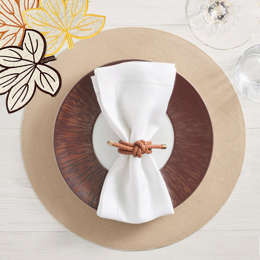 A table setting with a tan placemat, brown plate, and white napkin tied with the Kim Seybert Rope Knot Napkin Ring Set of 4 adds coastal charm. Wine and water glasses plus embroidered leaf details complete the look.
