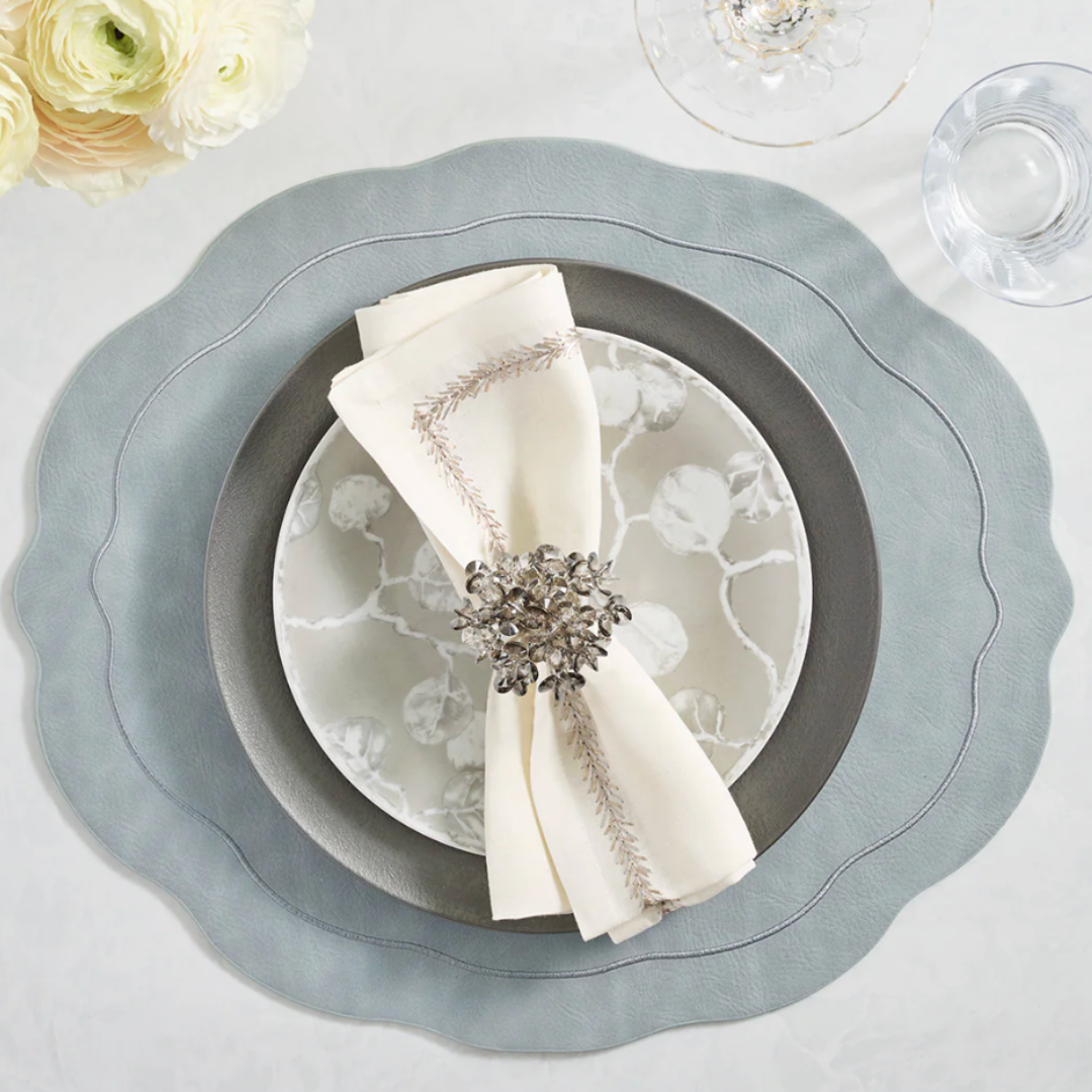 A formal place setting with a scalloped grey placemat, dark grey and white patterned plates, cream embroidered napkin, and Kim Seybert Aster Napkin Ring Set of 4, paired with wine and water glasses and elegant white flowers.