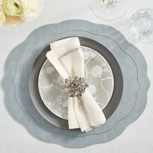 A formal place setting with a scalloped grey placemat, dark grey and white patterned plates, cream embroidered napkin, and Kim Seybert Aster Napkin Ring Set of 4, paired with wine and water glasses and elegant white flowers.