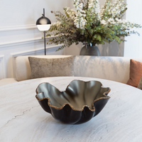 The Lirio R Matt Black & Bronze Bowl by Gardeco sits on a round marble table with a white sofa, neutral cushions, a modern lamp, and a tall floral arrangement in the background.