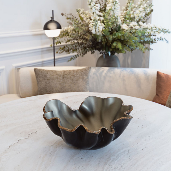 The Lirio R Matt Black & Bronze Bowl by Gardeco sits on a round marble table with a white sofa, neutral cushions, a modern lamp, and a tall floral arrangement in the background.