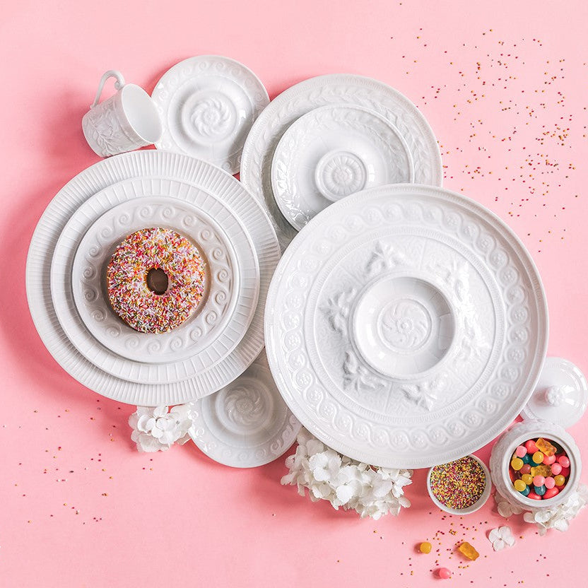 Bernardaud’s Louvre Dinnerware—service plates, bowls, and a mug—rests on a pink surface with colorful sprinkles and white flowers nearby. A sprinkled donut atop one plate adds playful charm to the elegant arrangement.