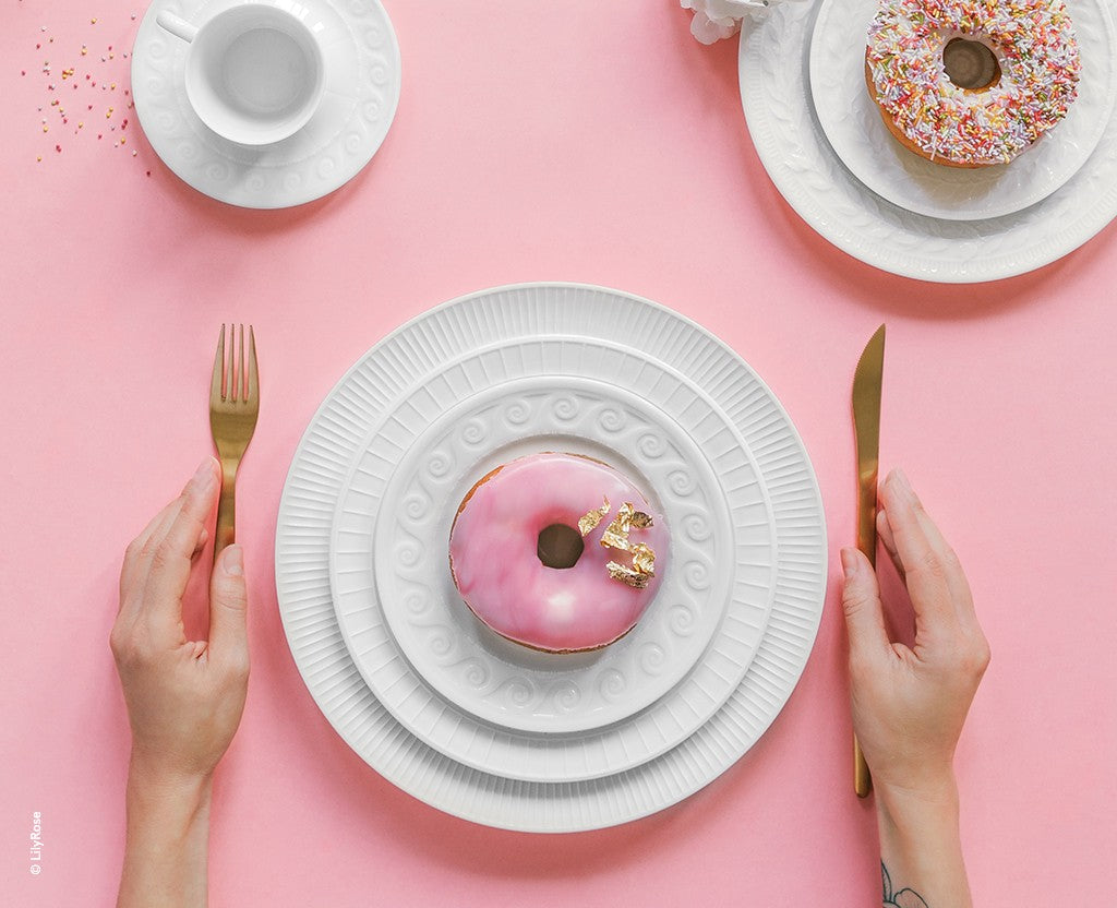 A person holds a gold fork and knife next to a pink donut with gold flakes on stacked Bernardaud Louvre Dinnerware plates, set against a pink background. Another Louvre plate with a sprinkle donut and a teacup are also shown.