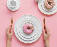 A person holds a gold fork and knife next to a pink donut with gold flakes on stacked Bernardaud Louvre Dinnerware plates, set against a pink background. Another Louvre plate with a sprinkle donut and a teacup are also shown.