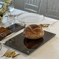 A round loaf of rustic bread sits on the BT-Shalom Lucite and Glass Wood Challah Board atop a white tablecloth, surrounded by gold cutlery, elegant plates, and flowers—perfect for a modern Shabbat gathering.