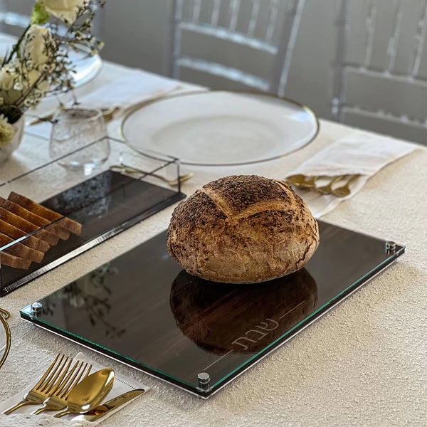 A round loaf of rustic bread sits on the BT-Shalom Lucite and Glass Wood Challah Board atop a white tablecloth, surrounded by gold cutlery, elegant plates, and flowers—perfect for a modern Shabbat gathering.
