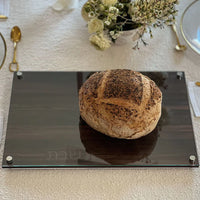 A round rustic loaf rests on the BT-Shalom Lucite and Glass Wood Challah Board, surrounded by elegant tableware and white flowers on a white tablecloth—ideal for a stylish Shabbat setting.