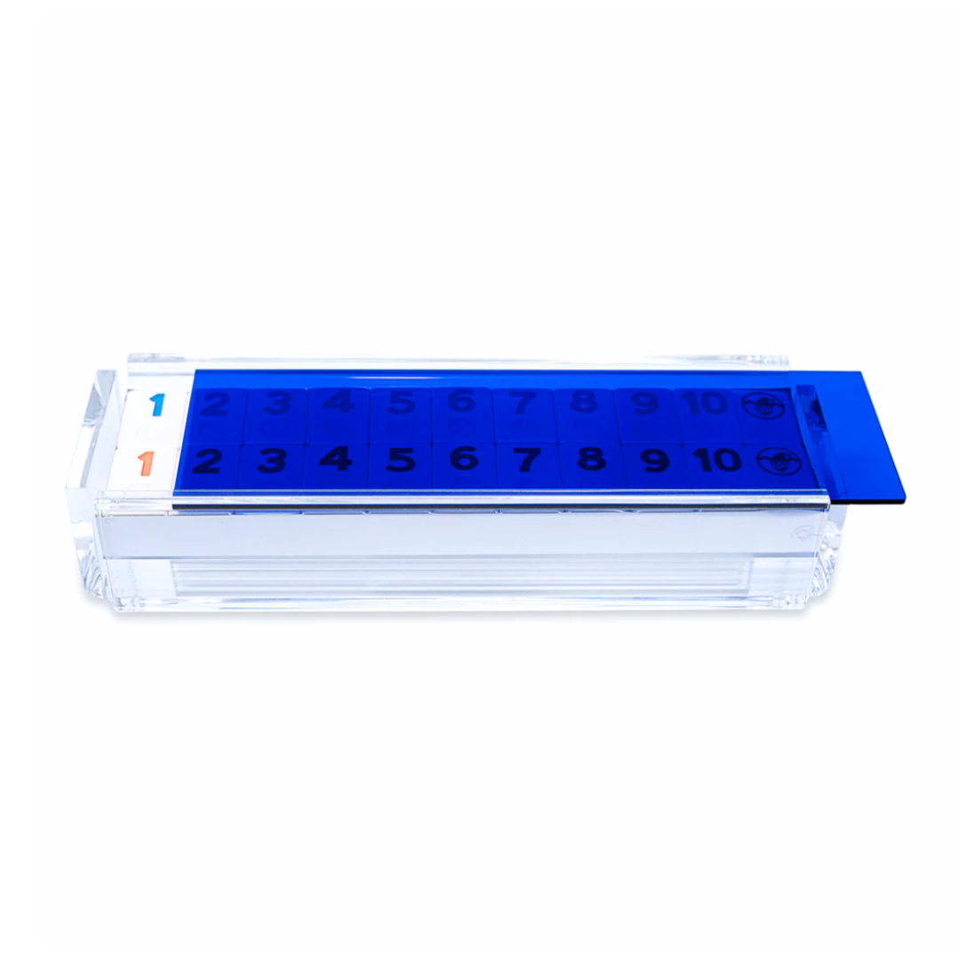 The Luxe Dominoes Rummy Acrylic Game Set features a clear rectangular case with a sliding blue lid, black numbers 1-10 on top, and a left sticker displaying blue and orange 1’s—a unique gift for any rummikub fan.