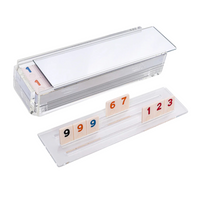 The Luxe Dominoes Rummy Acrylic Game Set features a clear holder with numbered tiles in red, blue, and black—one closed with tiles inside, the other displaying 9, 9, 6, 7, 1, 2, 3—making it an ideal gift.