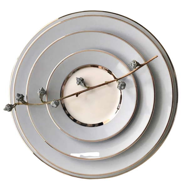 Four stacked Recamier MAK Grey & Platinum plates by Royal Limoges/Bia Cordon Bleu are arranged from largest to smallest, with a delicate branch bearing small buds placed diagonally across this elegant dinnerware set.