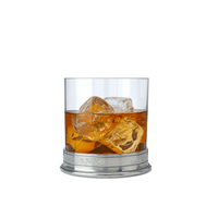 A glass of whiskey with large ice cubes sits on a MATCH Pewter Barware metallic base by MATCH, engraved with the initials JSM, set against a plain white background.