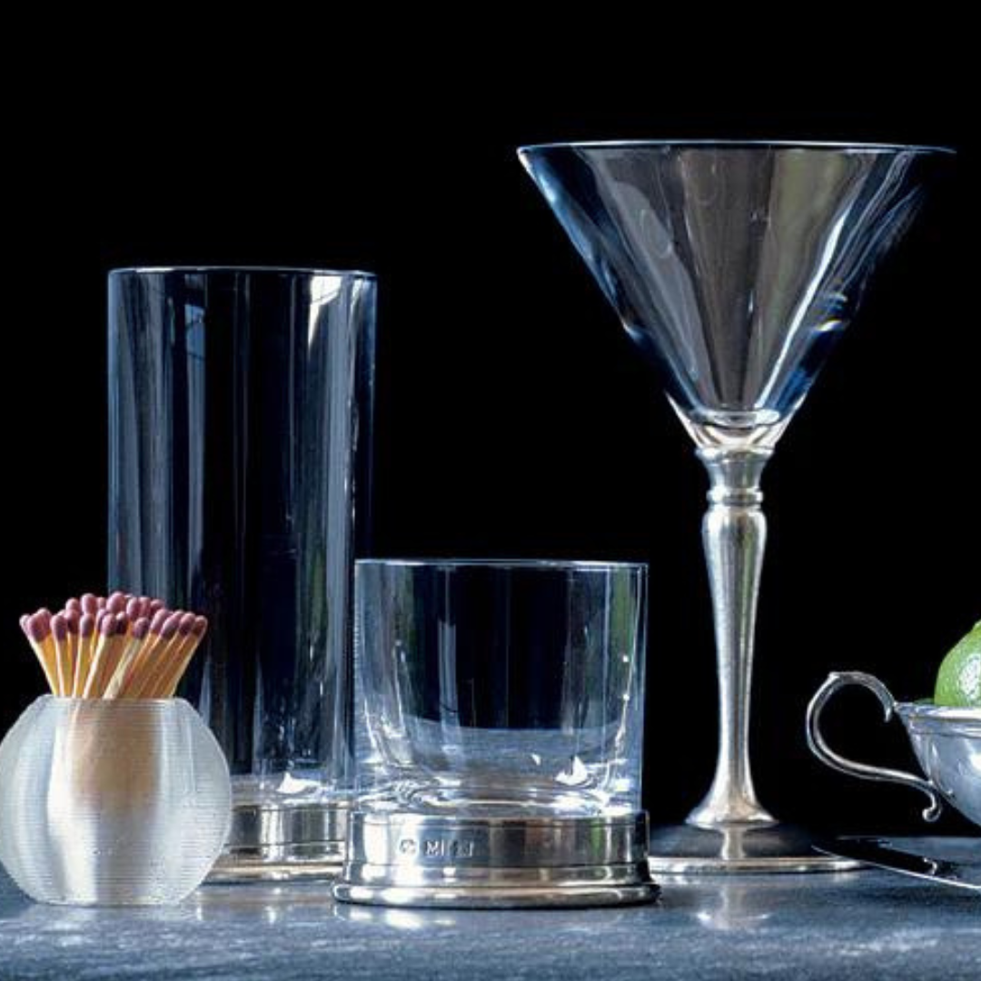 MATCH Pewter Barware by MATCH, including lead-free European crystal glasses (tall, short, martini) and Italian pewter accents—a match holder and metal dish with lime—are arranged on a dark surface.