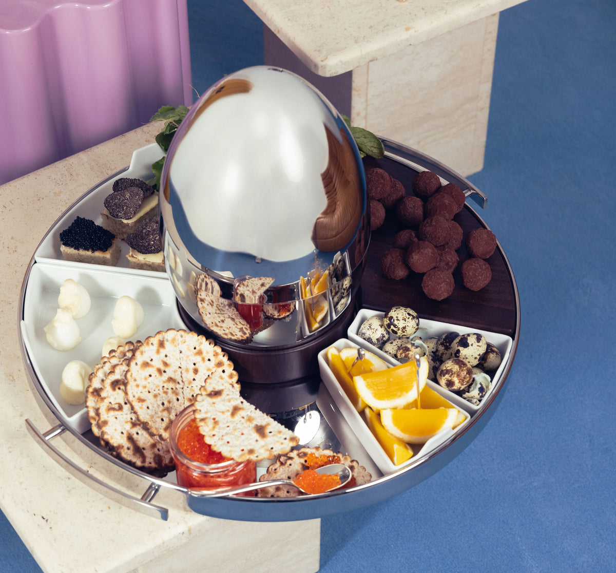 A round Christofle MOOD PARTY Stainless Steel Round Tray 15.7″ with a central dome, containing compartments filled with caviar, crackers, quail eggs, orange slices, and chocolate truffles.