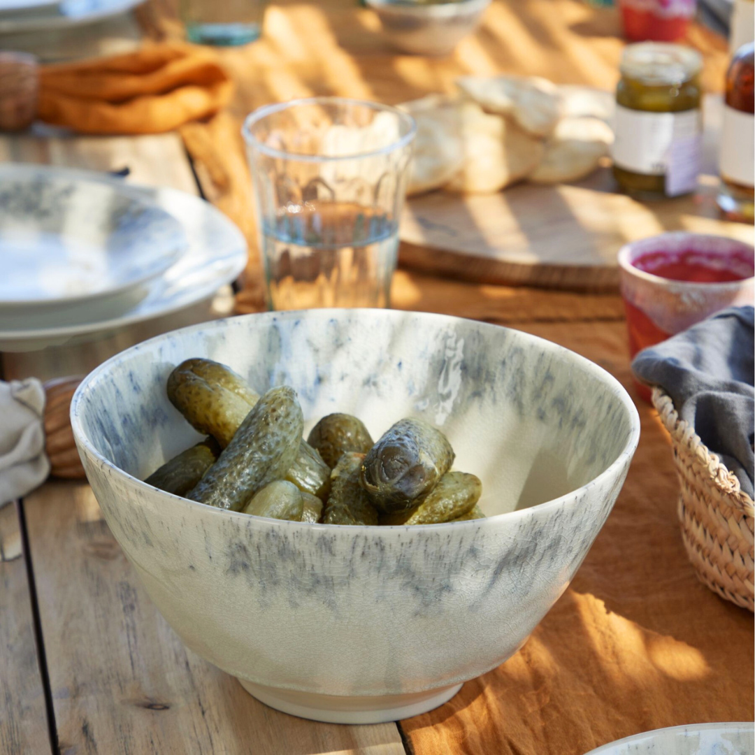 A Costa Nova Madeira Dinnerware Cream bowl filled with pickles sits on a wooden table set for a meal, surrounded by plates, glasses, bread, and small dishes under natural sunlight.