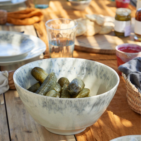 A Costa Nova Madeira Dinnerware Cream bowl filled with pickles sits on a wooden table set for a meal, surrounded by plates, glasses, bread, and small dishes under natural sunlight.