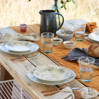 A rustic outdoor table set features Costa Nova's Madeira Dinnerware Cream with patterned plates, bowls of food, clear glasses of water, cutlery, a loaf of bread, a black pitcher on a mustard runner, and a vase with greenery for added charm.