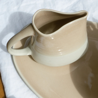 The Maguelone Dinnerware Tamaris ceramic pitcher by Jars - Rosenthal sits on its matching plate atop white fabric, with sunlight casting gentle shadows, highlighting its beige and cream crackled glazes.