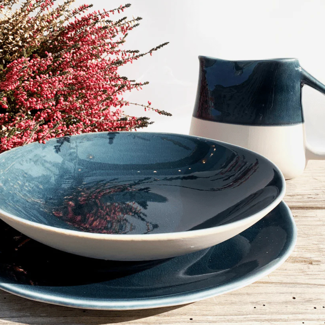 A Maguelone Outremer Dark Blue ceramic bowl and plate set by Jars - Rosenthal rests on a wooden surface beside a matching pitcher and pink heather flowers, all bathed in bright natural light.
