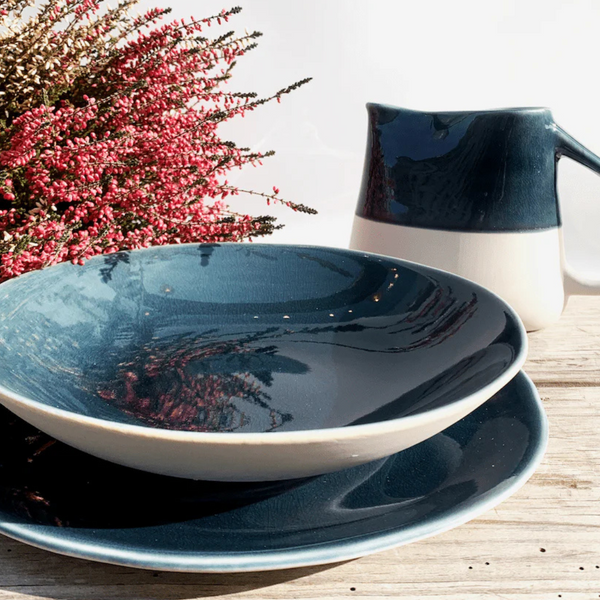 A Maguelone Outremer Dark Blue ceramic bowl and plate set by Jars - Rosenthal rests on a wooden surface beside a matching pitcher and pink heather flowers, all bathed in bright natural light.