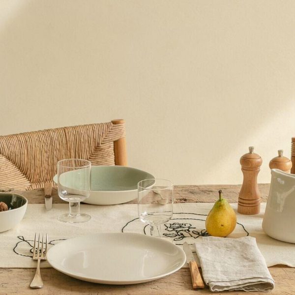 A rustic table features Maguelone Dinnerware Quartz by Jars - Rosenthal, a fork, knife, glasses, a pear, a green ceramic serving bowl, and wooden salt and pepper grinders on a runner with embroidered flowers.